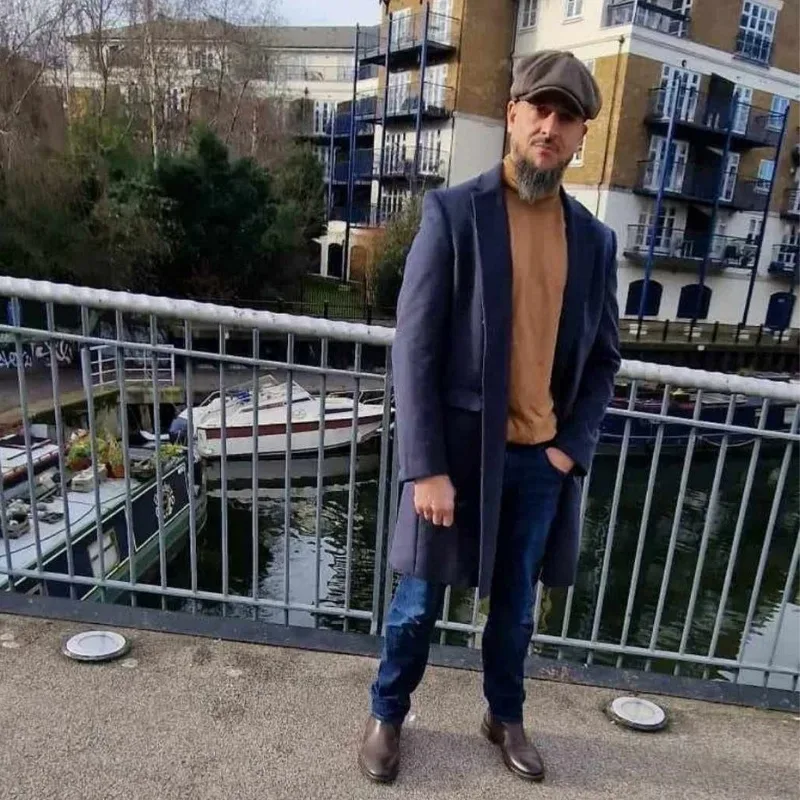 A man wearing a hat and a trench coat stands on a bridge overlooking a canal.