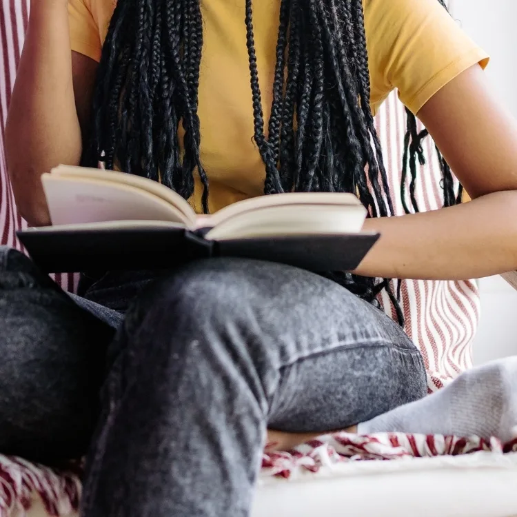 A person with long braids, wearing a yellow top and jeans, sits in a chair while reading a book near a bright window